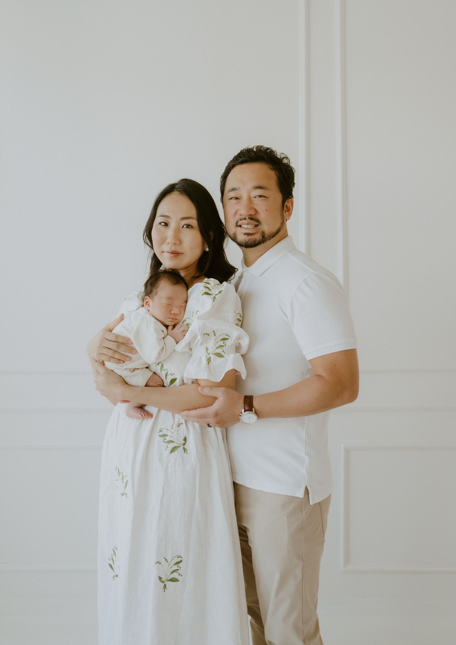 Family portrait with couple in white clothing holding baby against white wall.