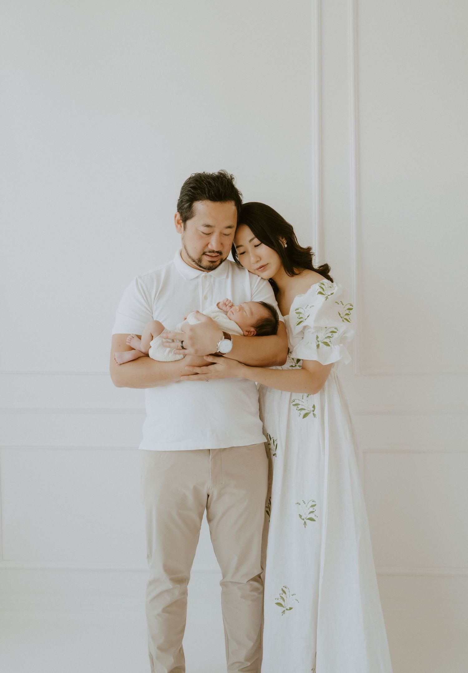 Parents sharing intimate moment with sleeping newborn in white room.