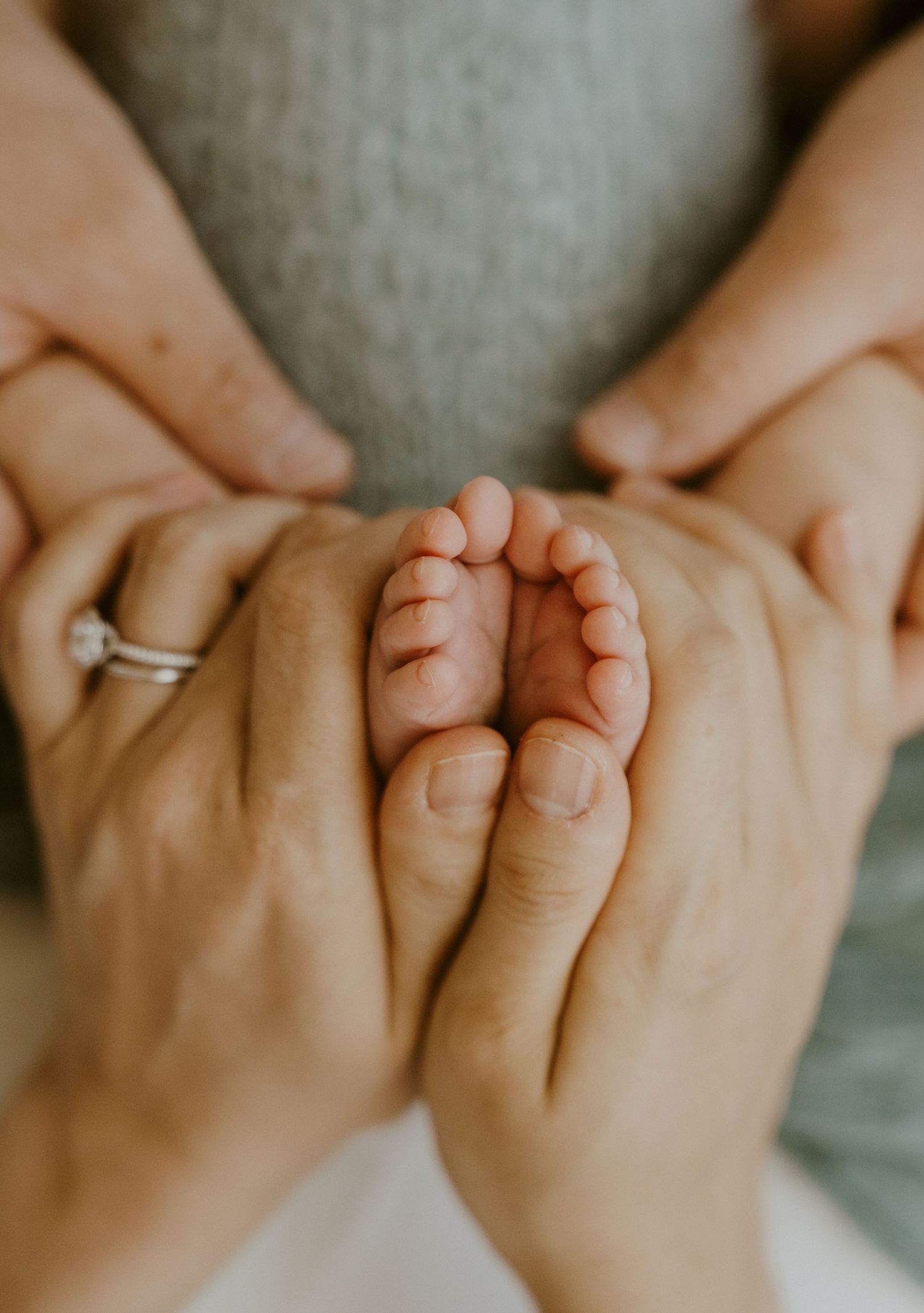 Adult hands gently holding tiny baby feet with engagement ring visible against gray background.