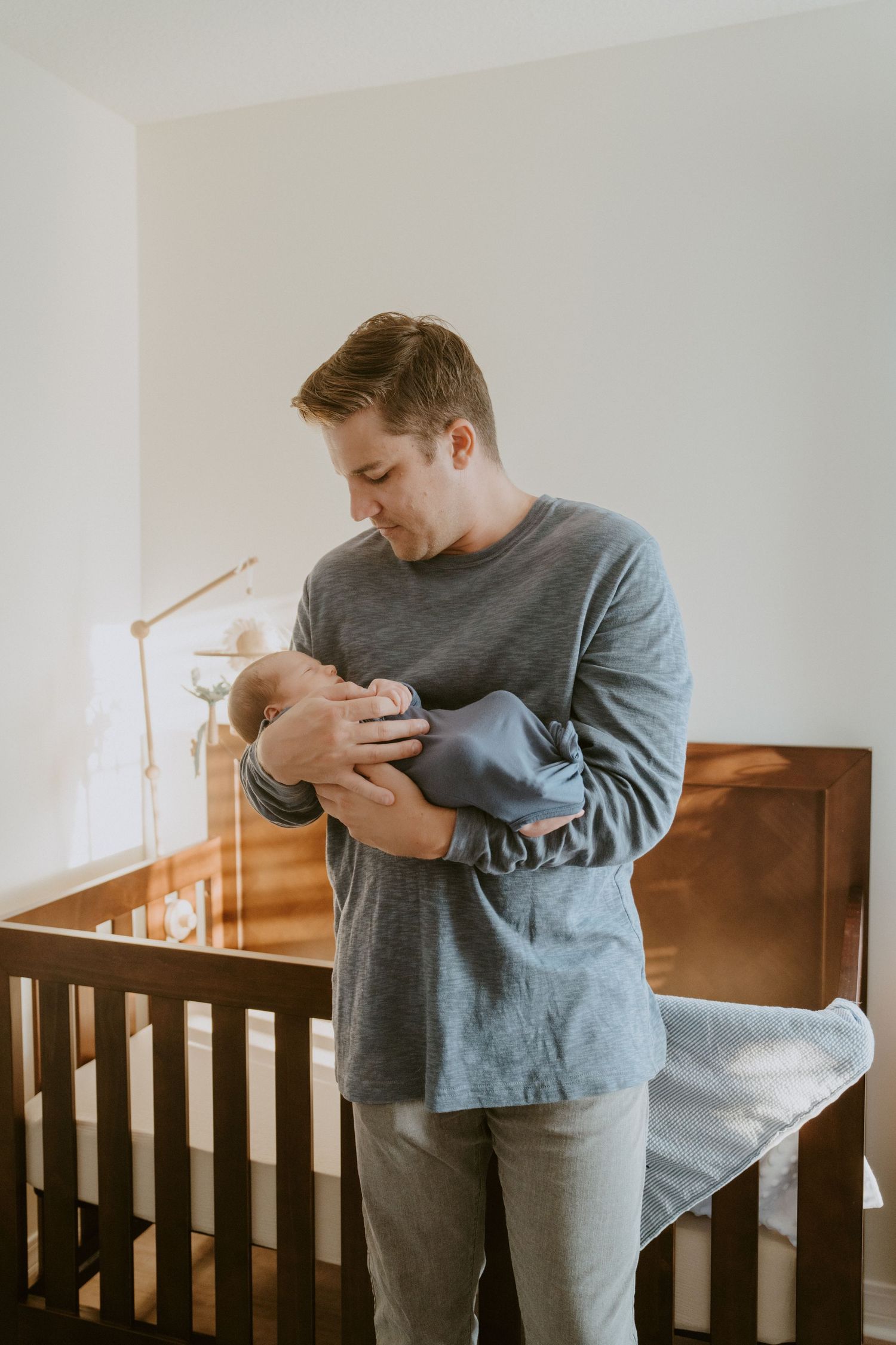 Parent holding newborn baby beside wooden crib in nursery with soft lighting.