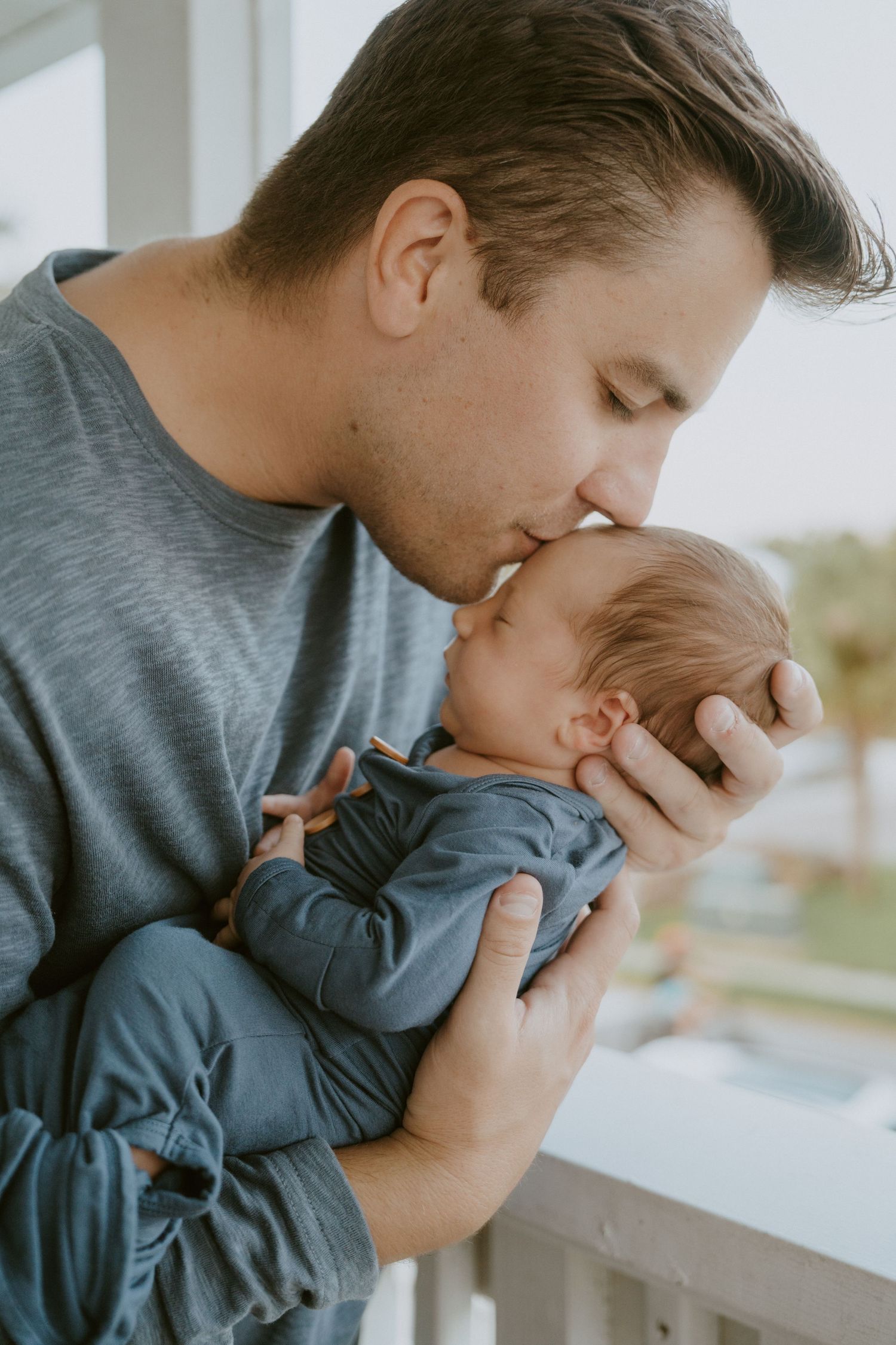 Parent giving gentle kiss to newborn baby near window in soft natural light.