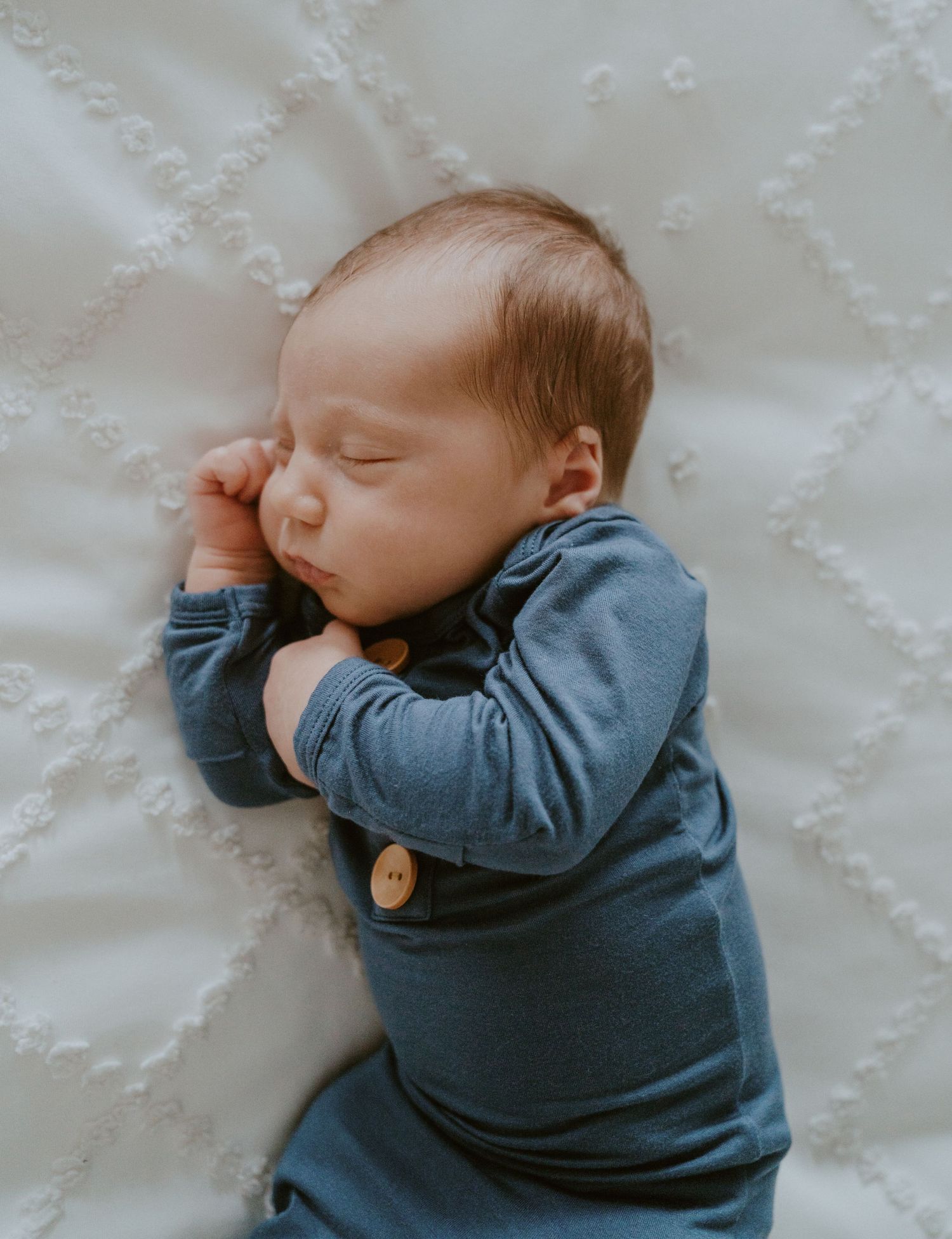 Sleeping newborn wearing blue outfit rests peacefully on white textured bedding.