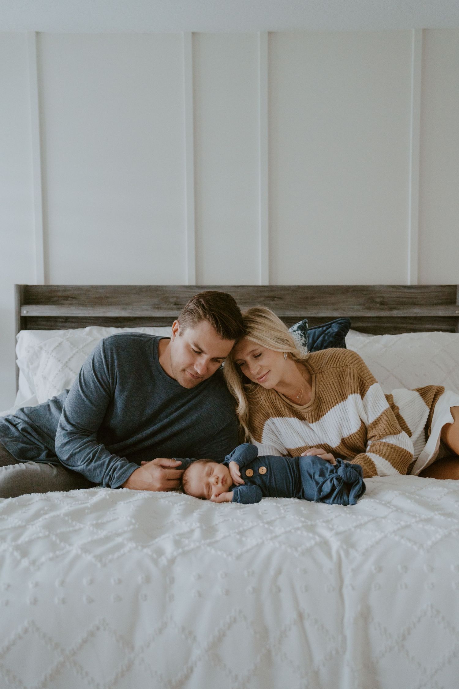 Family snuggling together on bed with white linens and wooden headboard.