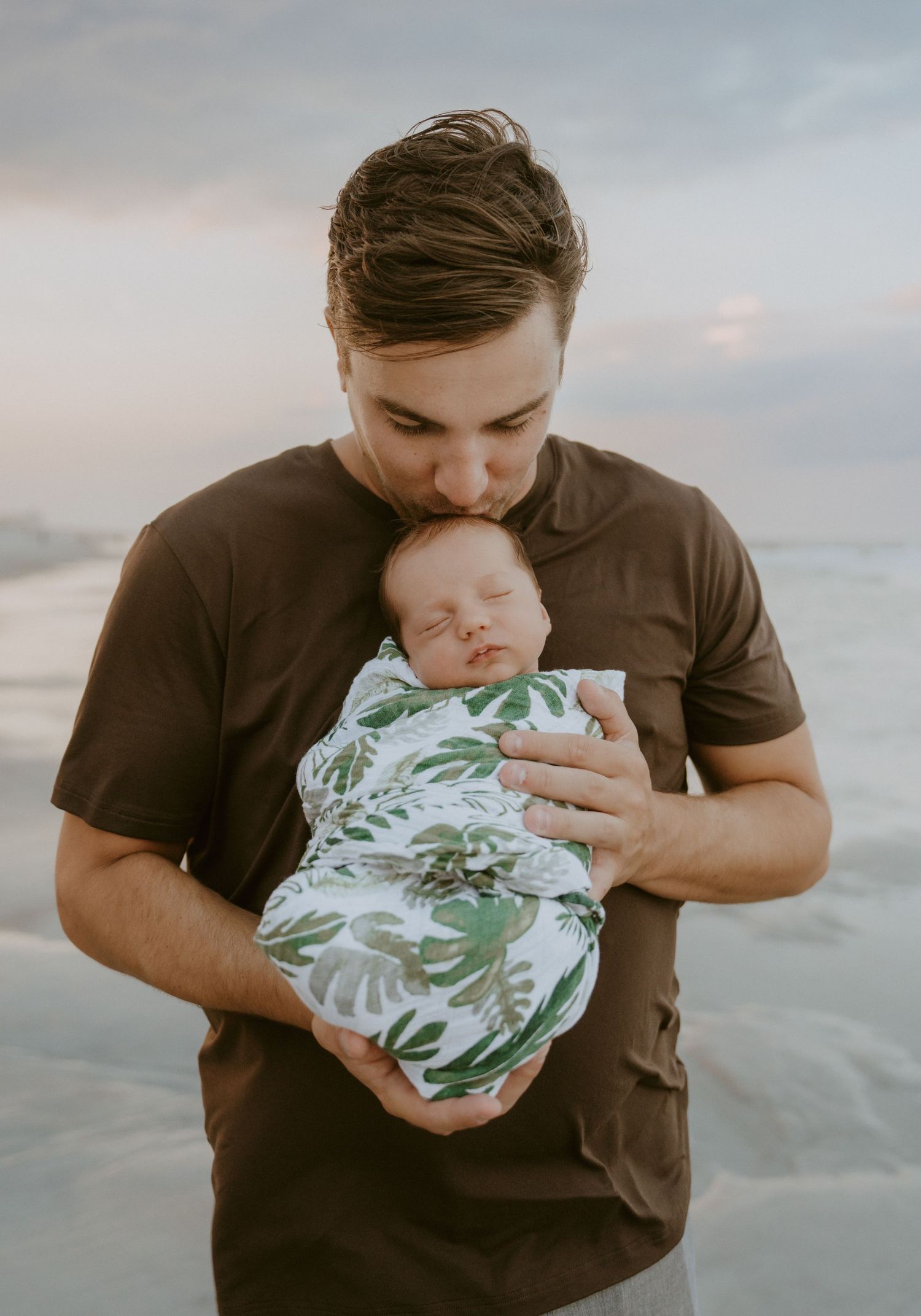 Parent holding sleeping infant wrapped in tropical print swaddle at sunset.