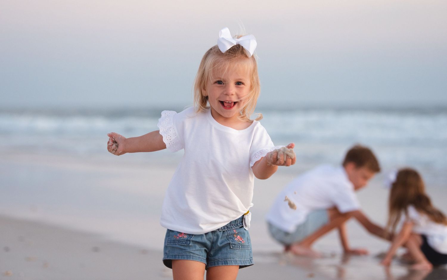 Young child in white top and denim shorts runs joyfully on beach as other children play in background.