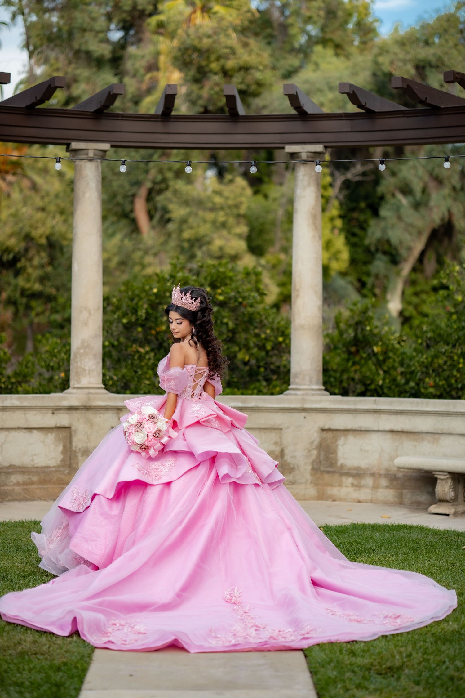 Young woman in a bright pink quinceañera ball gown with a crown poses under a pergola in a garden.