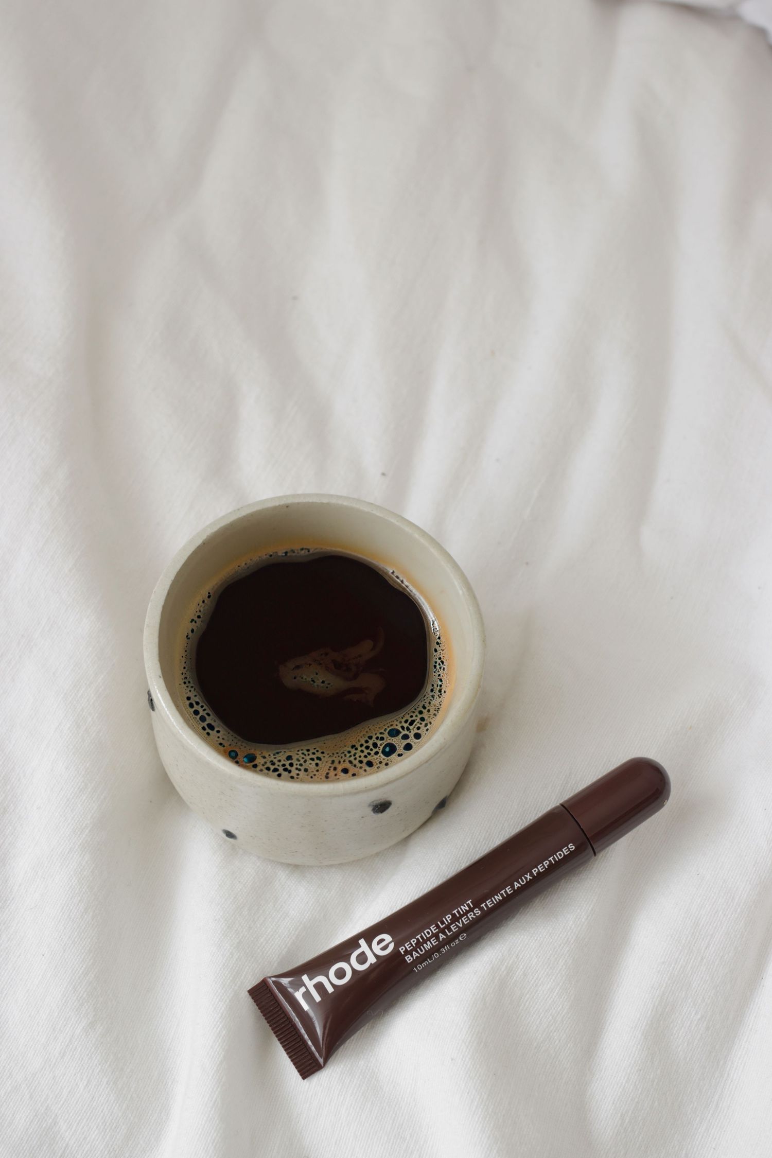 Coffee cup and brown cosmetic tube on white textile surface.