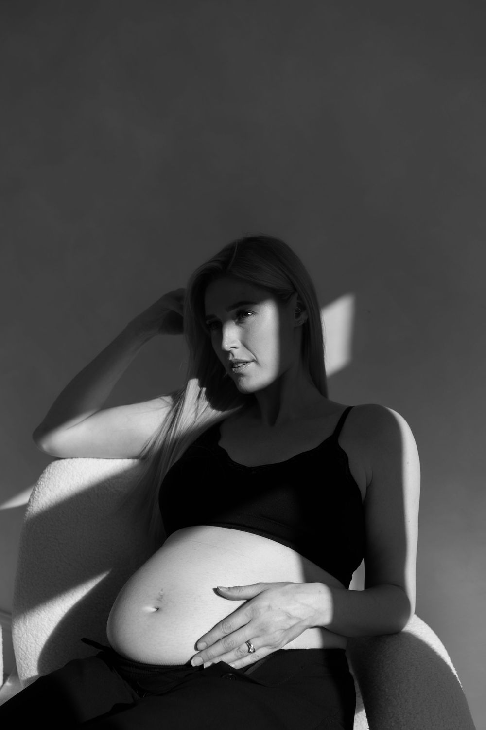 full black and white profile of woman sitting in boucle chair in a spot of sunlight showcasing her bump.