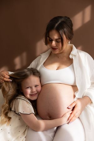 woman holding daughter while she hugs her mothers bump infant of brown backdrop during maternity session based in Billingham