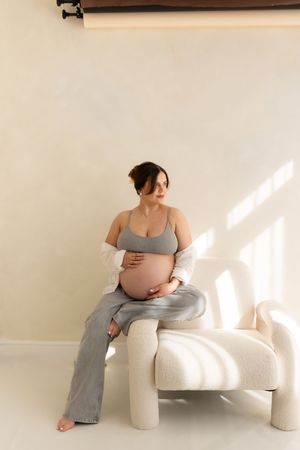 woman holding her bump in photography Teeside studio while sitting on the edge of a chair wearing jeans and a bra.