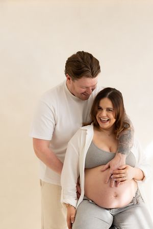 couple laughing together in the studio during a maternity session based in Zoe Cullen photography studio. Women is sitting on a stool.