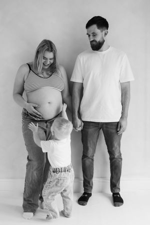 black and white image of couple stood against wall as little boy runs up to touch her bump during their maternity session