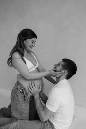 black and white photo of woman kneeling in between partners legs while holding her partners face as he holds her bump during maternity shoot