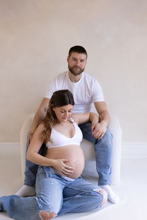 woman sitting in-between mans legs while he sits on boucle chair holding his hand and her bump during their maternity session