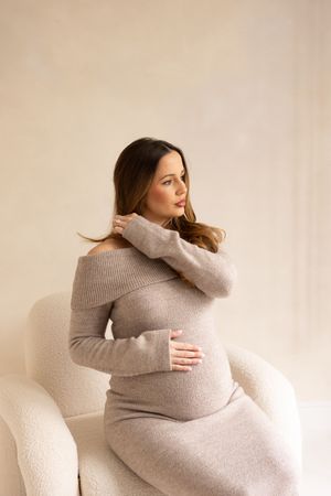 woman playing with her hair while sitting on boucle chair holding her bump during her maternity session in Northeast studio.