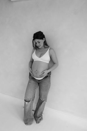 black and white photo of woman leaning against studio wall holding her bump during her maternity session in Teeside studio.