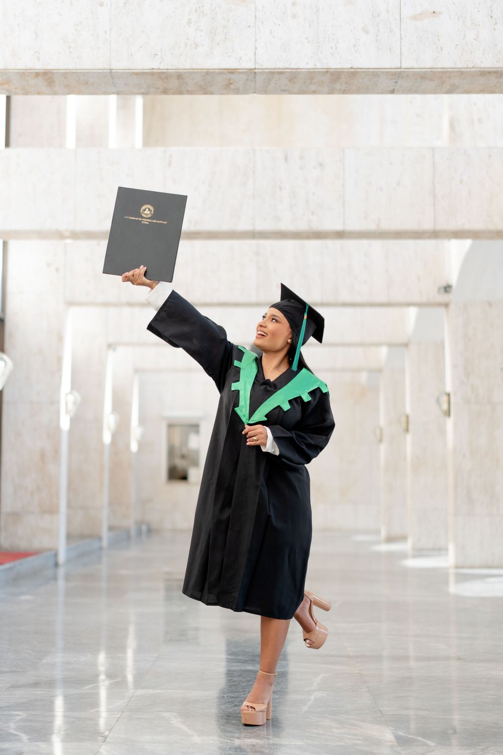 Graduate in cap and gown holding diploma triumphantly in bright hallway.