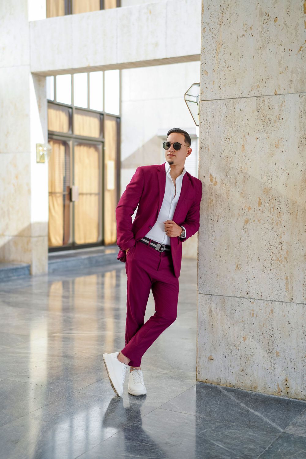 Stylish man in magenta suit and white sneakers poses against marble wall.