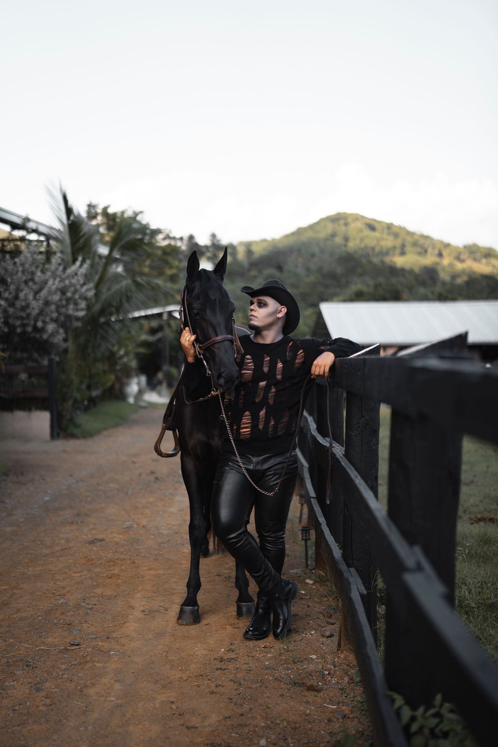 Equestrian in black attire poses with a dark horse on a dirt path near a lake at sunset.