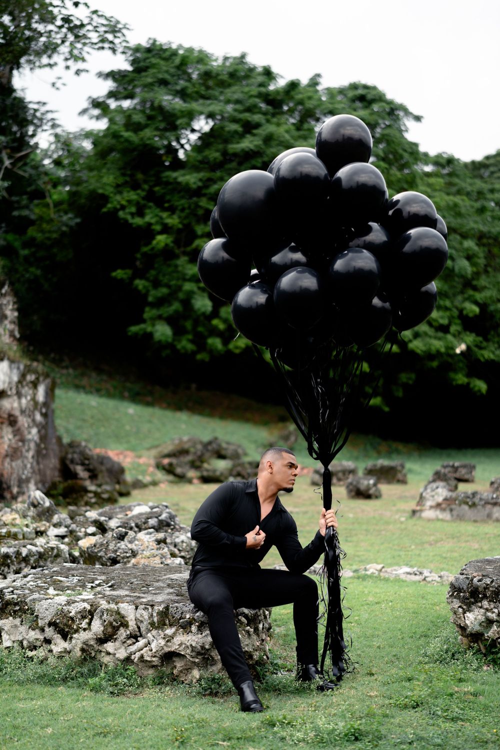 Person in black attire kneeling with bunch of black balloons in garden setting.