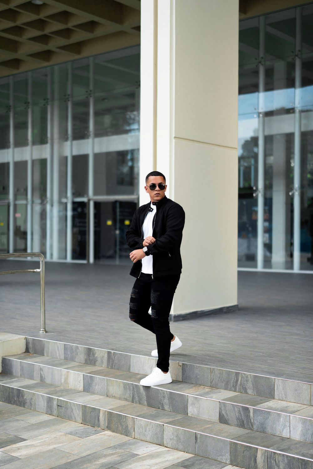 Man in black outfit and sunglasses poses on marble steps outside modern building.