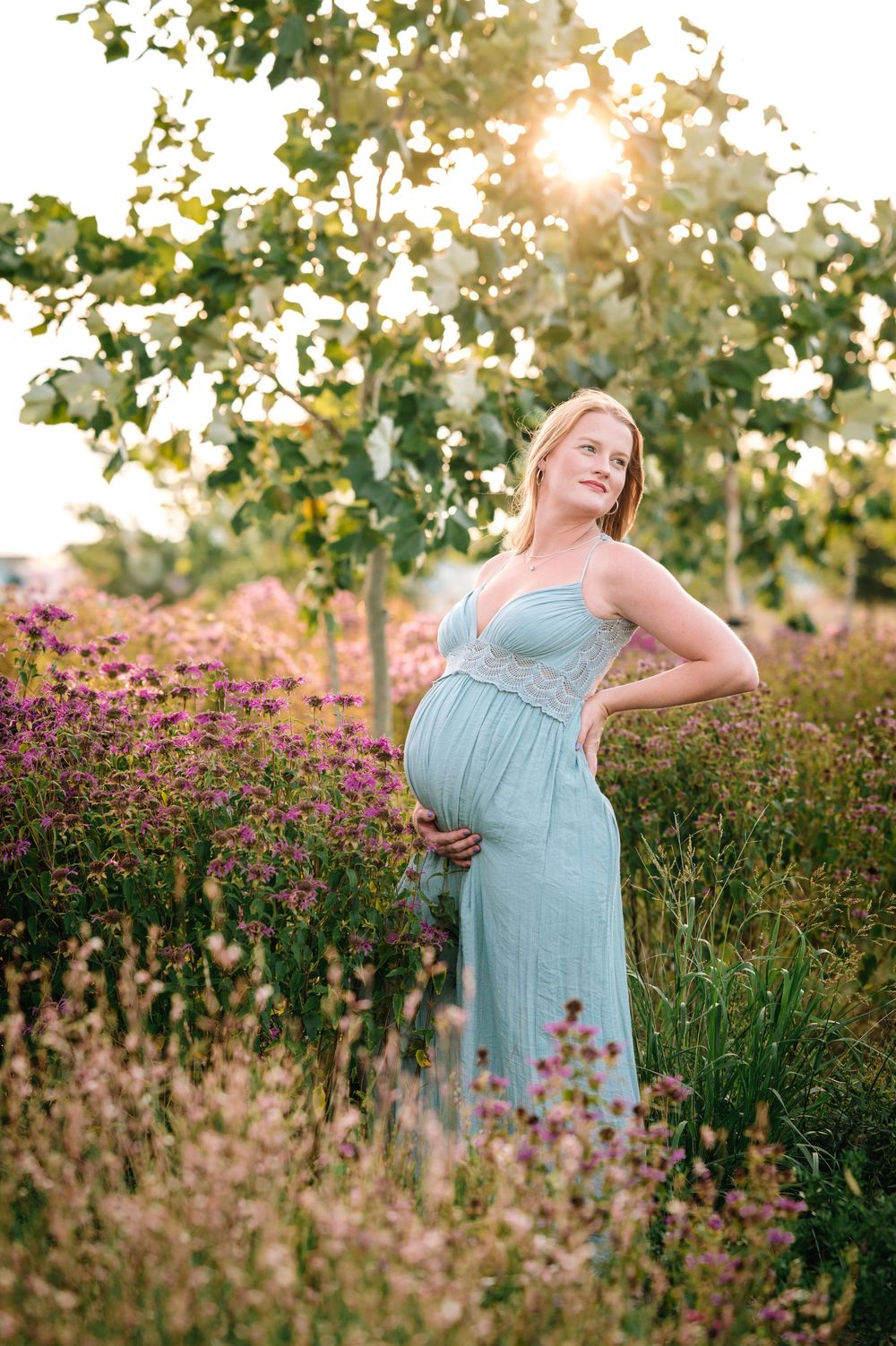 Expectant mother in turquoise dress poses in field of purple flowers at sunset.