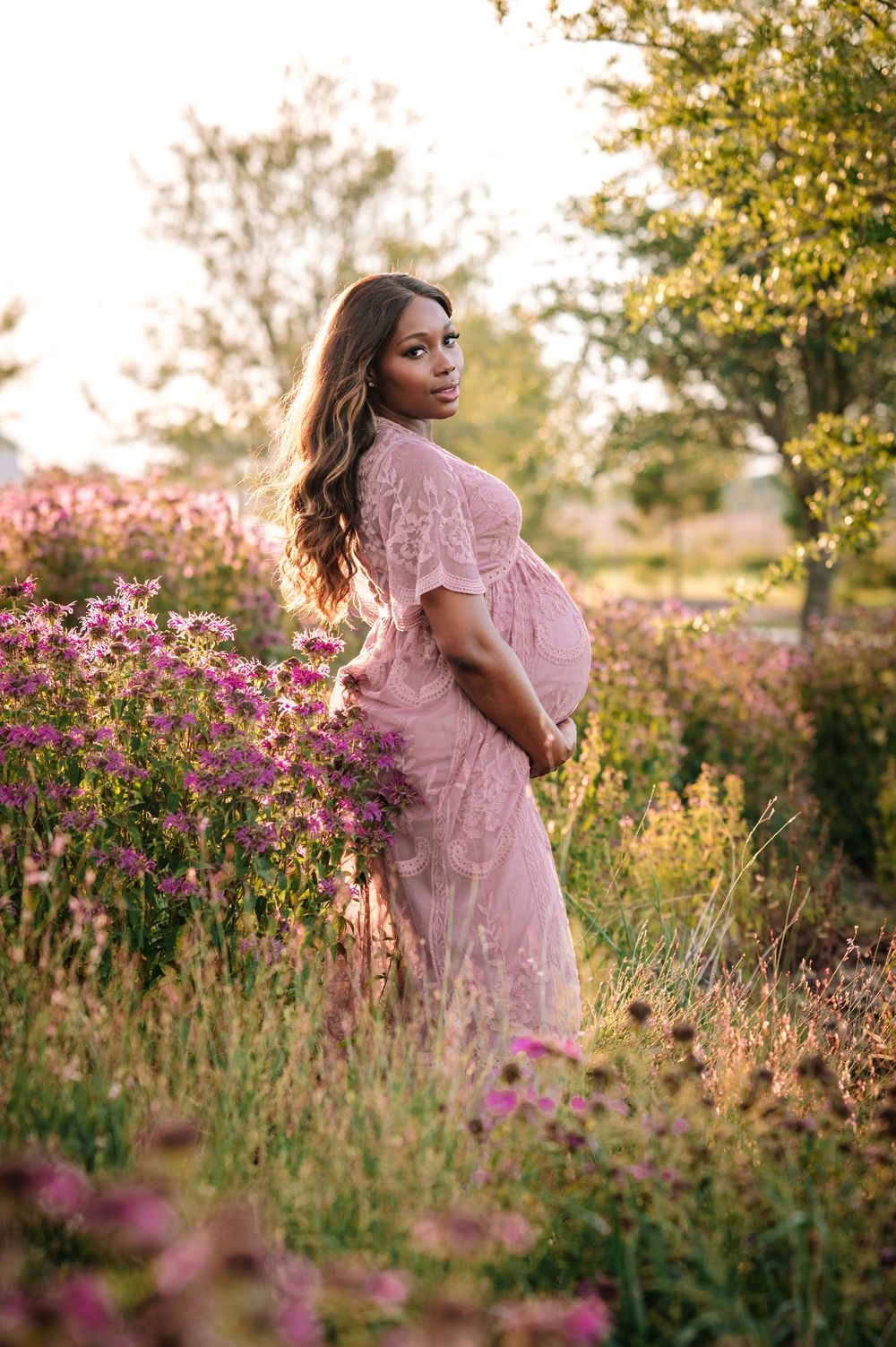 Golden hour maternity photo with woman in pink dress surrounded by purple wildflowers.