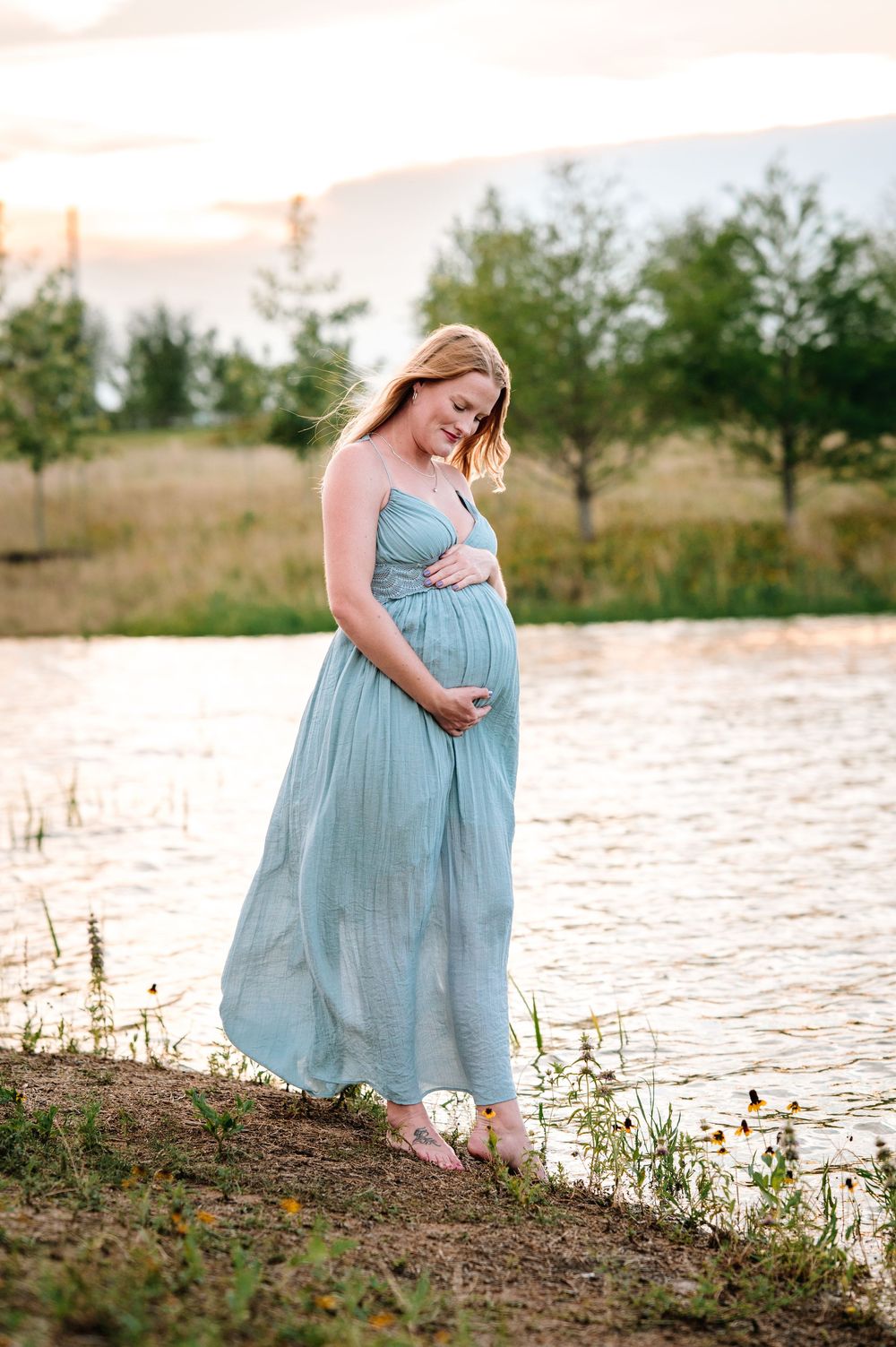 Pregnant woman in flowing blue dress walks along waterfront during sunset.