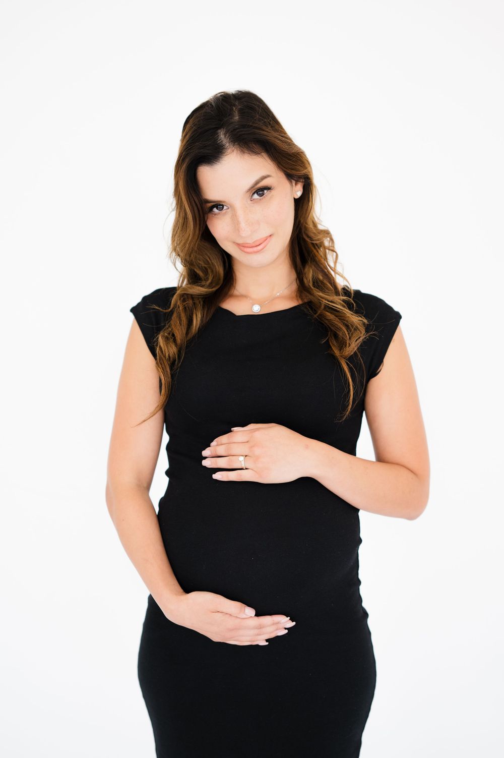 Expectant mother in black dress poses against white backdrop during maternity session.