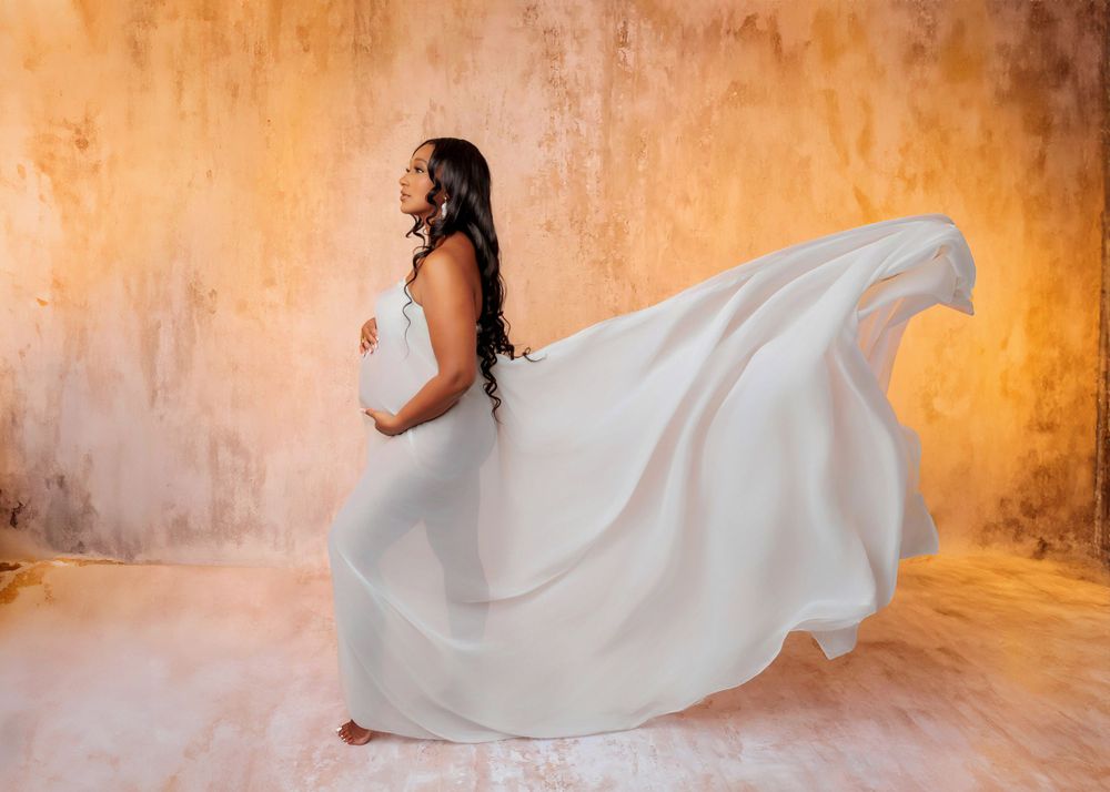 Pregnant woman in flowing white gown with billowing fabric poses against warm textured golden backdrop.