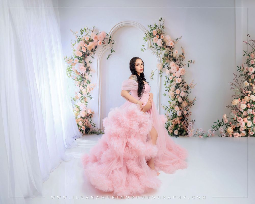 Pregnant woman in a pink fluffy tulle gown poses before a floral arch backdrop in a studio maternity shoot.