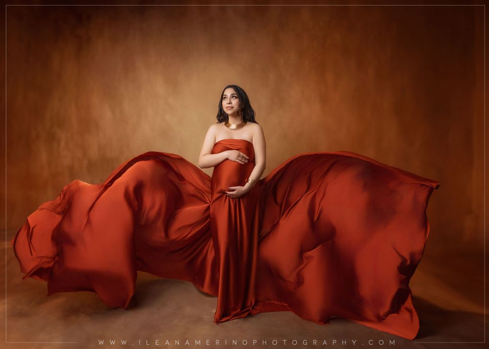 Pregnant woman in dramatic red silk flying gown poses gracefully against warm brown studio background.