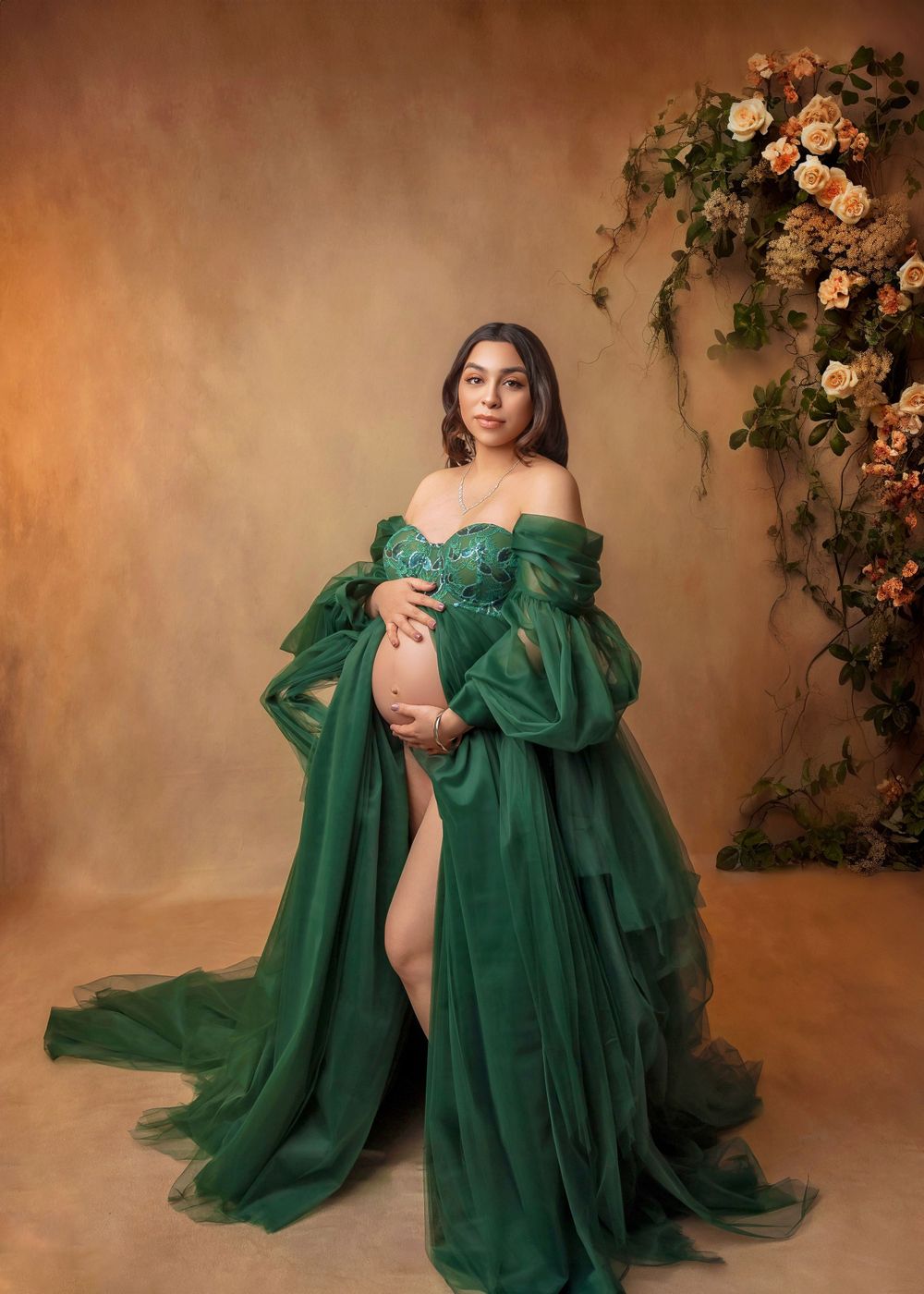 Expectant mother in emerald green tulle maternity robe poses beside floral arrangement on golden backdrop.