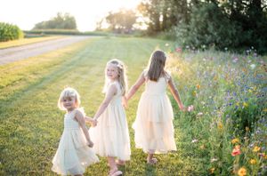 Three small figures in white dresses walk together through a wildflower field at golden hour.