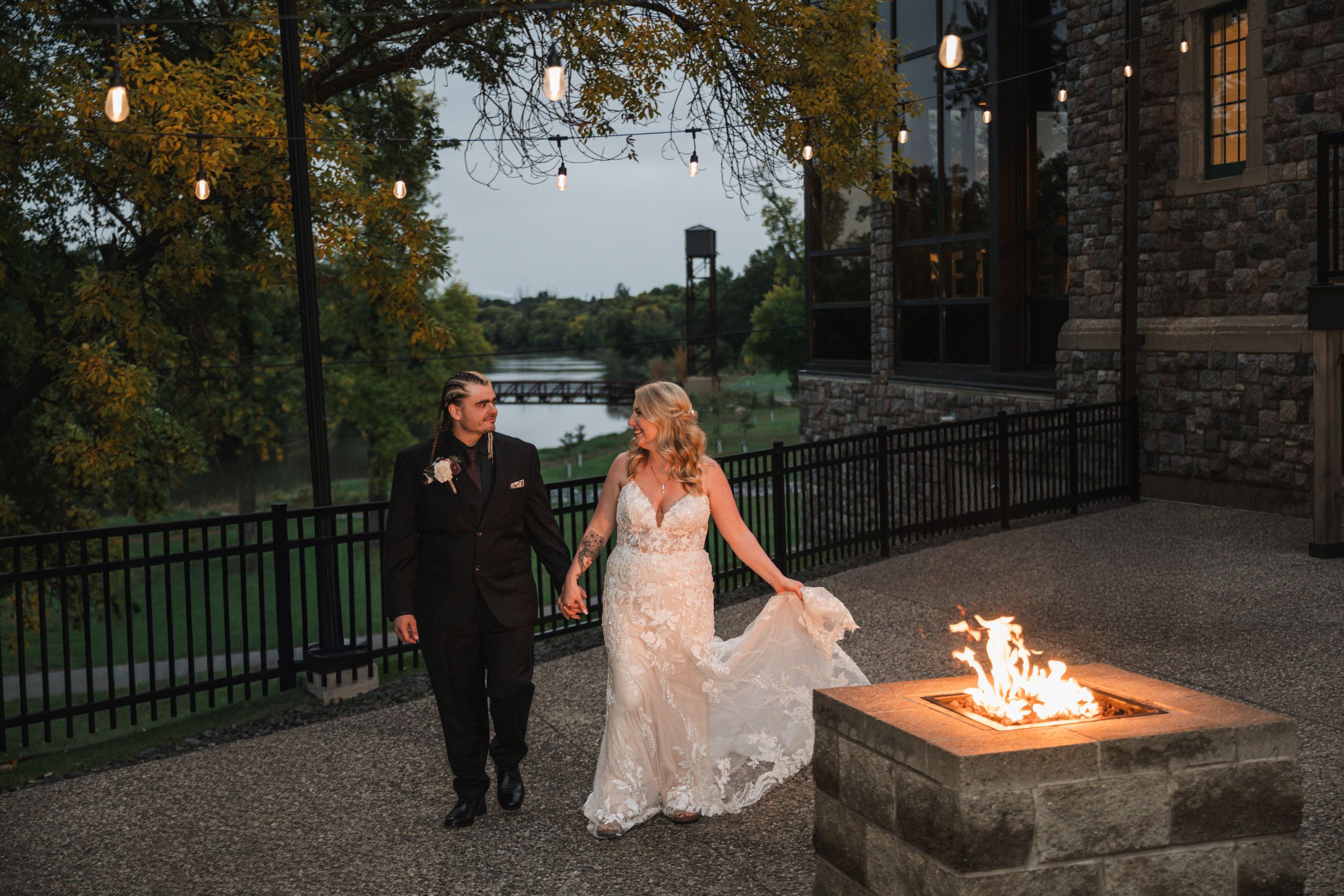 Bride and groom hold hands walking past a stone fire pit at an elegant outdoor wedding venue at dusk.