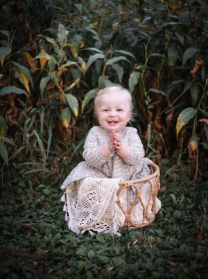 Smiling baby wearing vintage lace dress sits in wicker basket outdoors among foliage.