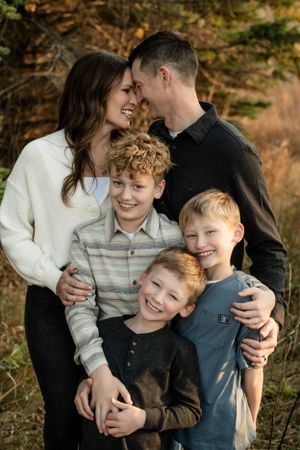 Family portrait outdoors with three young children against autumn forest background.