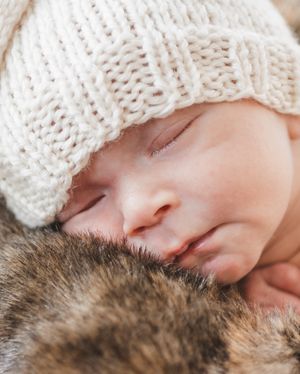 Sleeping newborn wearing white knit hat resting on fuzzy brown blanket.