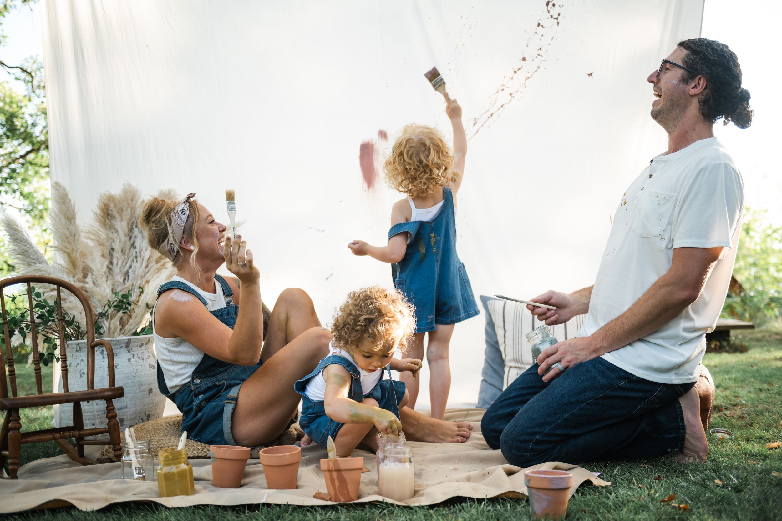 Family of four enjoying outdoor painting activity on grass, with children playing and parents smiling on a sunny day.