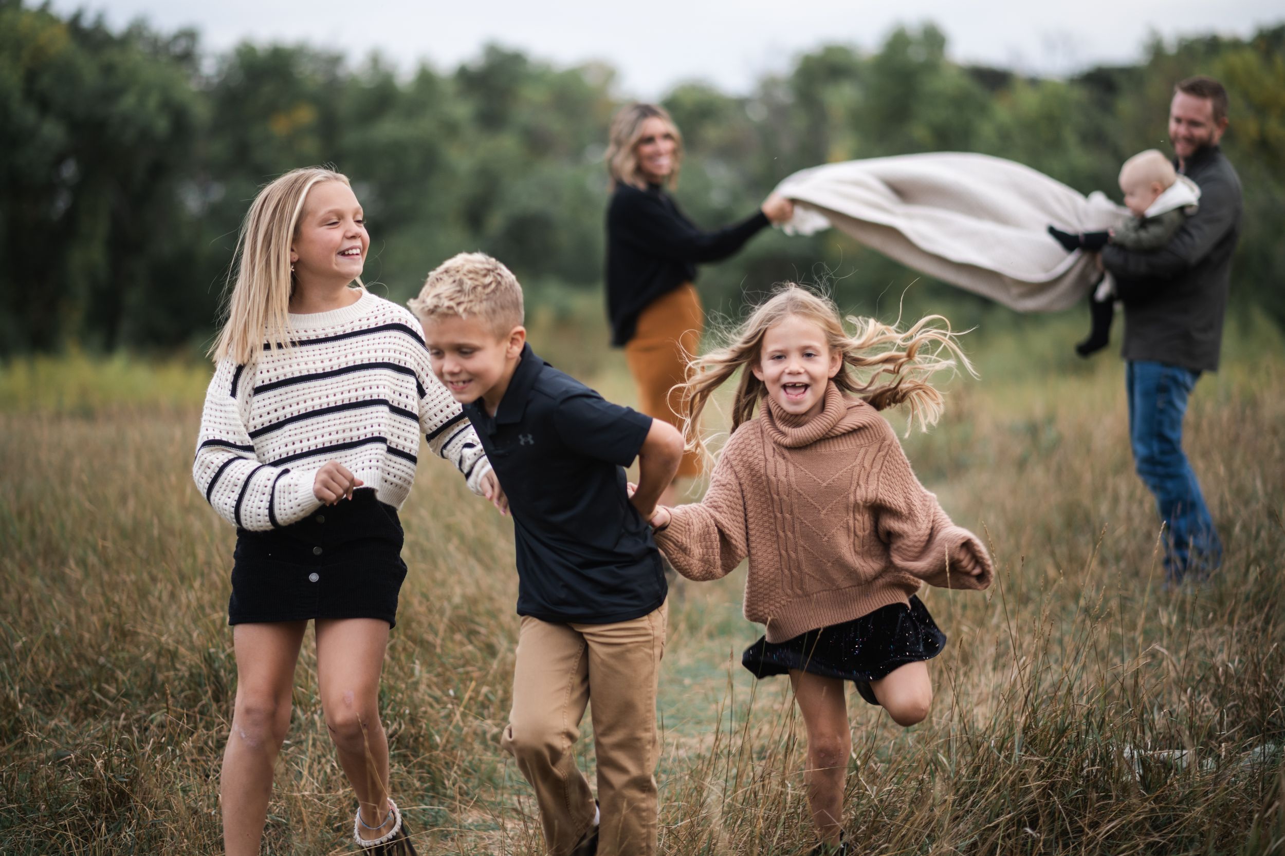 Happy family with three children running through a grassy field while parents play with a blanket in background.