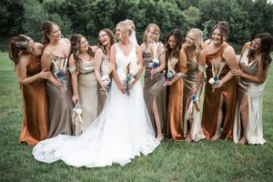 Wedding party wearing elegant mix of earth-toned dresses gather on green lawn for group photo.
