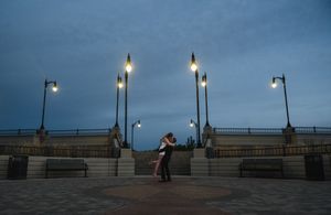 Couple embracing at dusk on a waterfront plaza with glowing lamp posts under a dramatic blue cloudy sky.