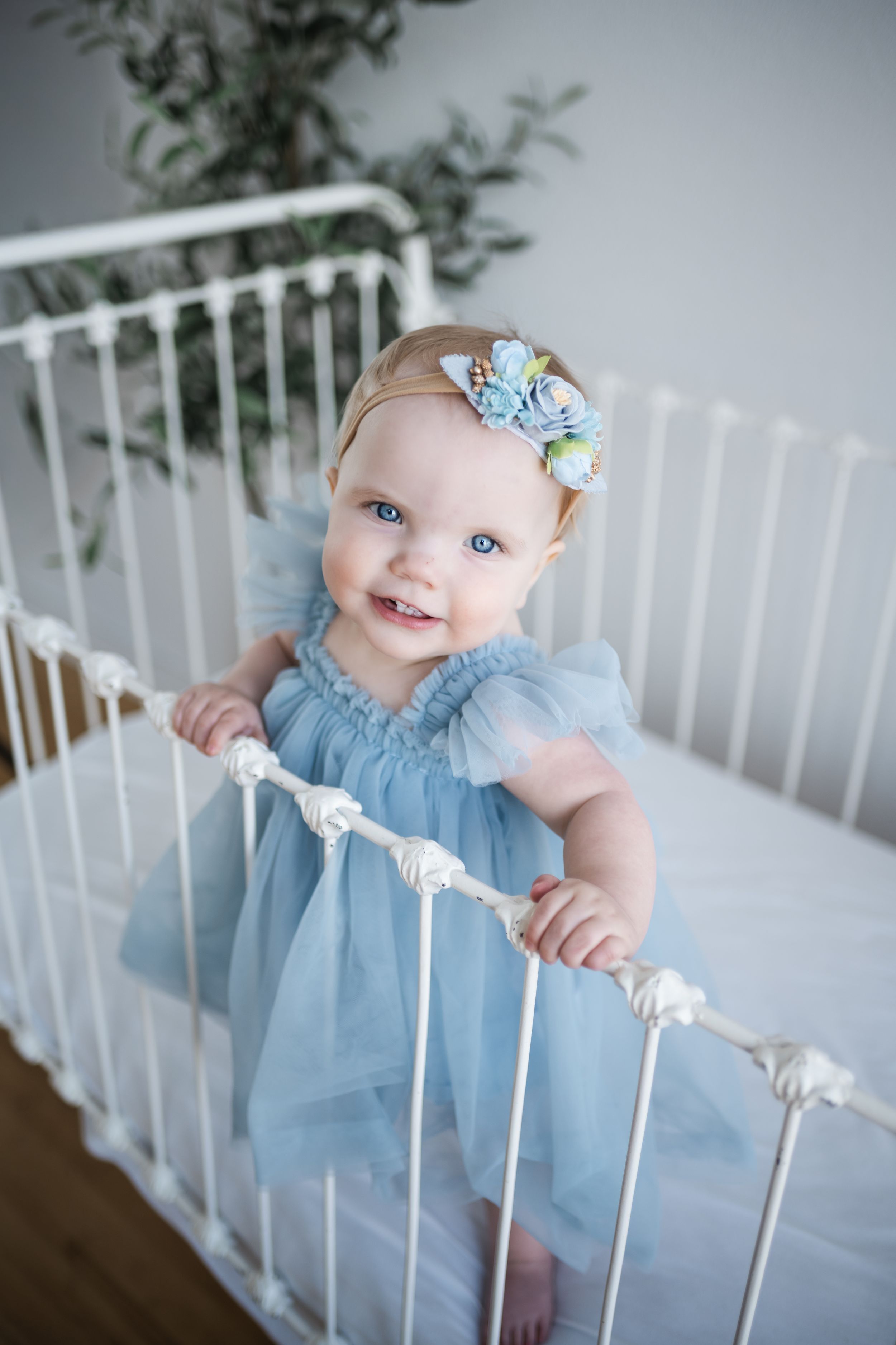 A baby in a light blue dress smiles while standing in a white crib with greenery in the background.
