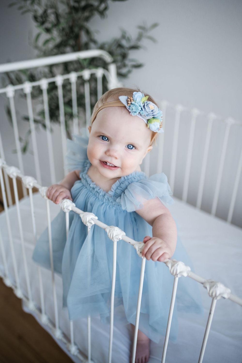 A baby in a light blue dress smiles while standing in a white crib with greenery in the background.
