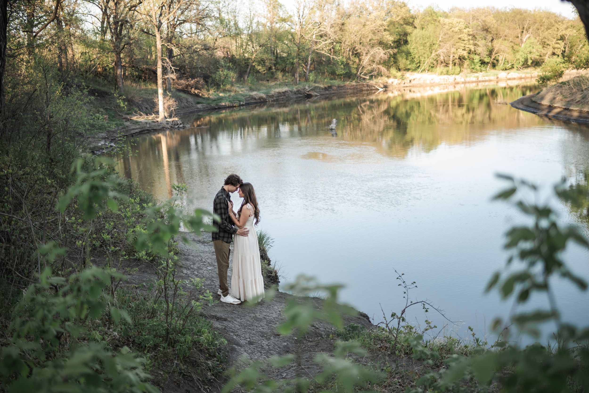 Couple embracing near a serene river surrounded by lush trees in a romantic outdoor wedding photo.