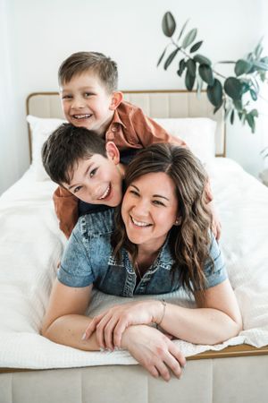 Playful family portrait on white bed with natural lighting and indoor plants.