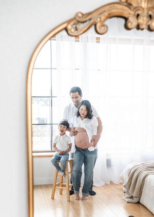 A reflection in an ornate gold mirror shows an intimate family moment in a bright bedroom.