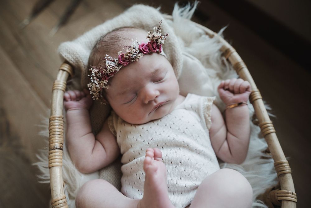 Sleeping newborn baby in floral crown headband resting in wicker basket with white fluffy blanket.
