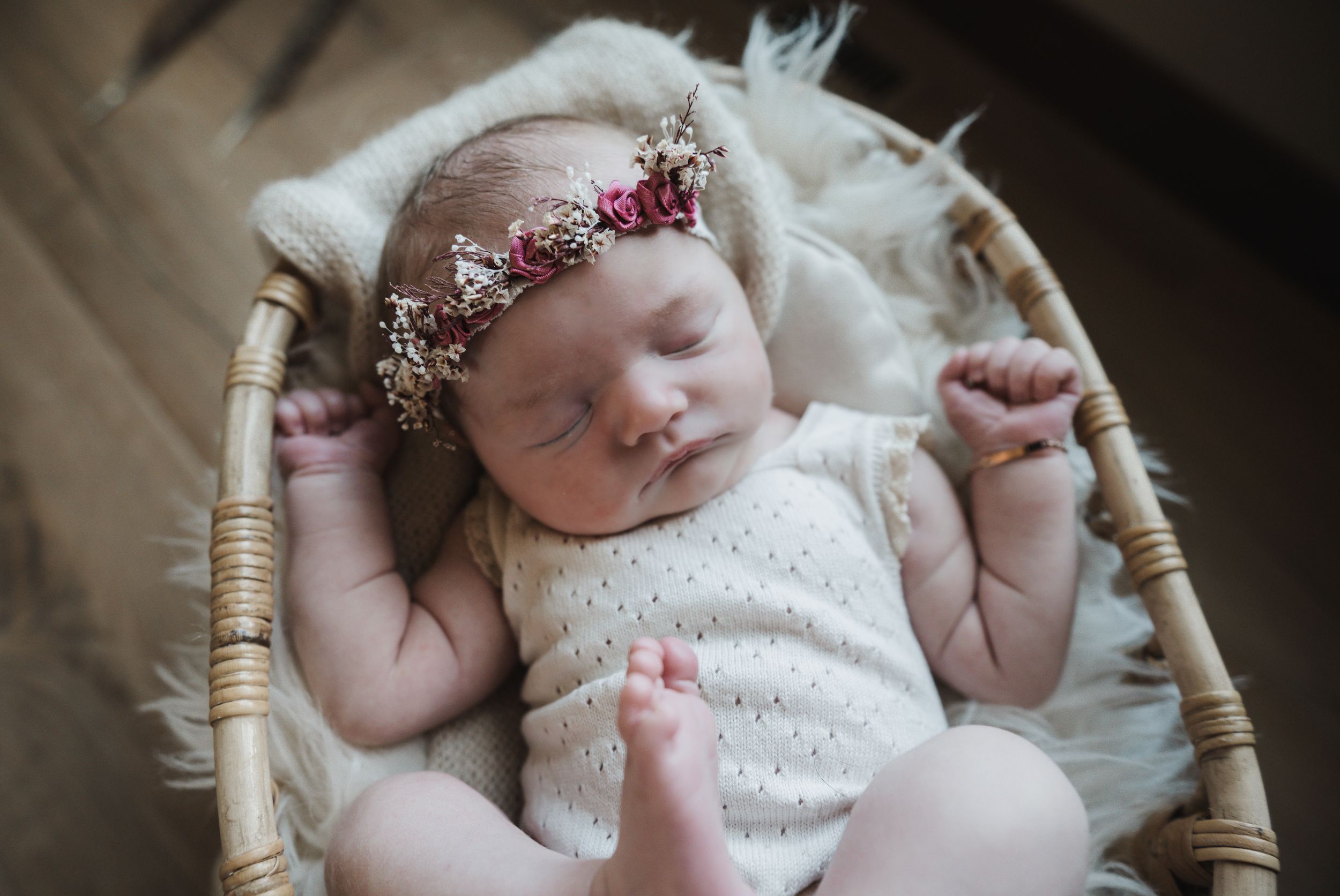 Sleeping newborn baby in floral crown headband resting in wicker basket with white fluffy blanket.