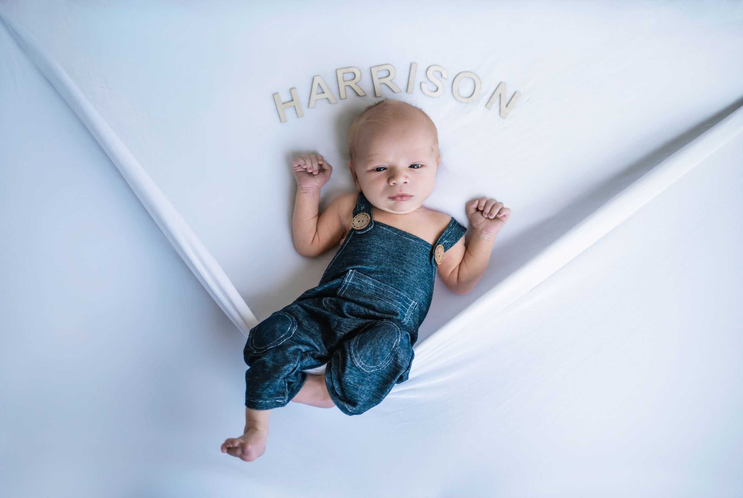 Baby Harrison in denim overalls lying on white fabric with name letters above, newborn portrait photography.